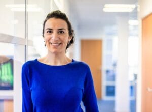 A photo of a person in an office setting smiling for a photo, they are wearing a blue jumper