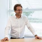 A portrait photo of a man in an office, he is wearing a white shirt and glasses and has both hands on a wooden desk