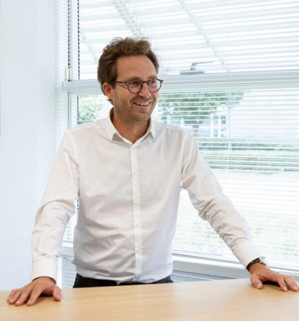 A portrait photo of a man in an office, he is wearing a white shirt and glasses and has both hands on a wooden desk