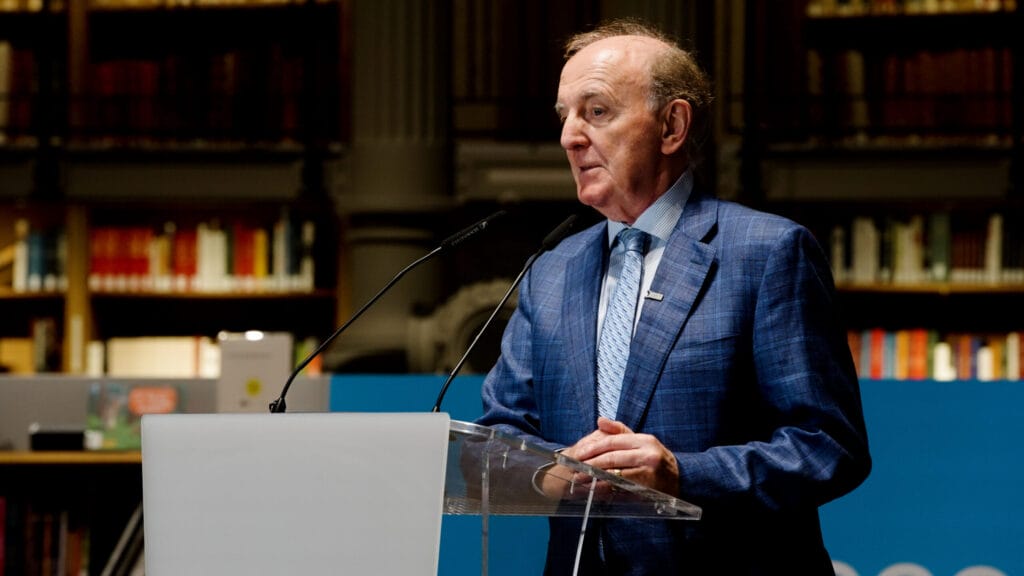 A man giving a speech at a podium, he is wearing a navy blue suit